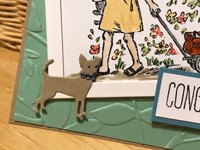 May include: A handmade greeting card featuring a die-cut tan dog with three small blue rhinestones on its collar. The card has a light green embossed leaf background and a white panel with a partial illustration of a person and the text CONG.