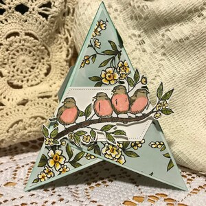 May include: A handmade, triangular greeting card featuring a branch with four birds and yellow flowers. The card has a light blue background with floral accents. The card is approximately 13 cm tall.