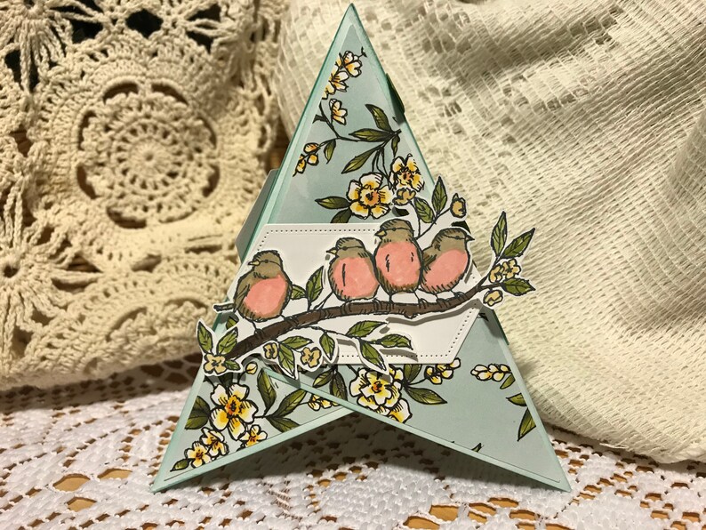May include: A handmade, triangular greeting card featuring a branch with four birds and yellow flowers. The card has a light blue background with floral accents. The card is approximately 13 cm tall.