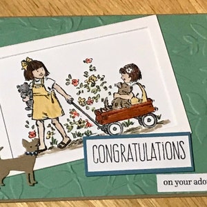 May include: A greeting card featuring a watercolor illustration of two children, one pulling a red wagon with a dog inside and the other holding a cat. The card includes the text CONGRATULATIONS on your adoption and a small brown dog cutout.