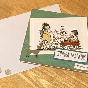 May include: A greeting card with a green embossed leaf pattern background featuring a hand-colored illustration of a child pulling a red wagon with another child and a dog inside. The card includes a brown dog cutout and text reading CONGRATULATIONS on your adoption, with a matching white envelope featuring two small paw prints.