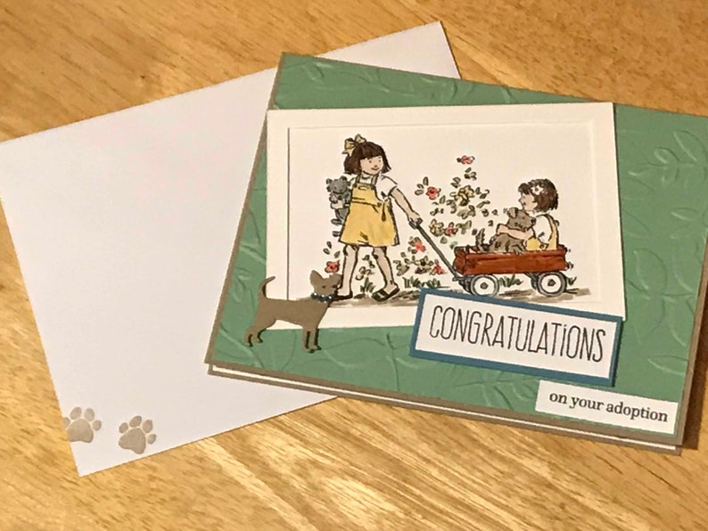 May include: A greeting card with a green embossed leaf pattern background featuring a hand-colored illustration of a child pulling a red wagon with another child and a dog inside. The card includes a brown dog cutout and text reading CONGRATULATIONS on your adoption, with a matching white envelope featuring two small paw prints.