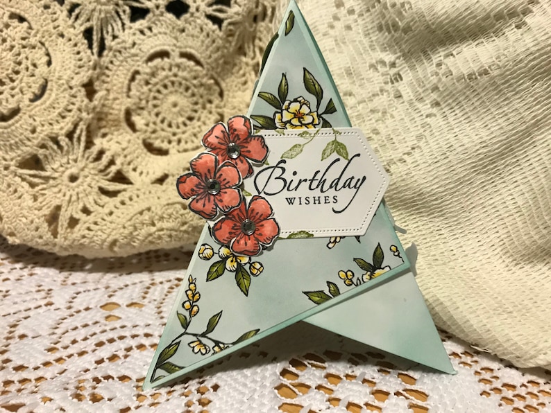 May include: A handmade birthday card in a triangular shape. The card is light blue with a floral pattern and features the words "Birthday Wishes" on a white label. It is decorated with orange flowers and green leaves.