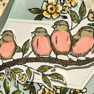 May include: A handmade card with a design of four birds on a branch. The birds have pink chests and brown and grey feathers. Green leaves and yellow flowers surround the birds. The card has a light blue background.