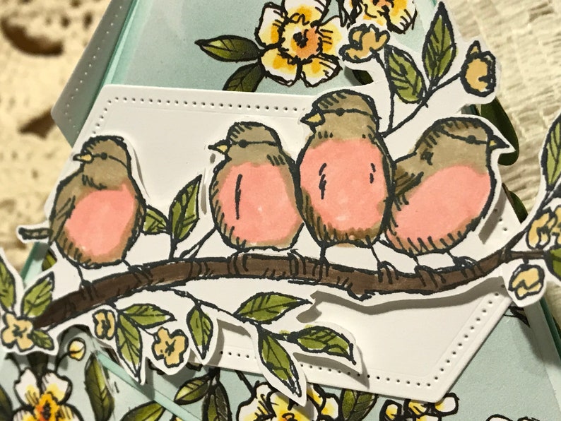 May include: A handmade card with a design of four birds on a branch. The birds have pink chests and brown and grey feathers. Green leaves and yellow flowers surround the birds. The card has a light blue background.