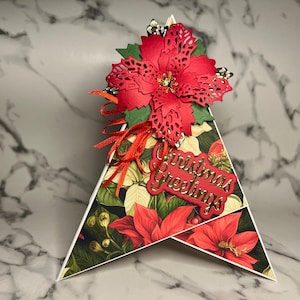 May include: A red and green Christmas card shaped like a tree with a poinsettia flower on top and the words "Christmas Greetings" in gold lettering.