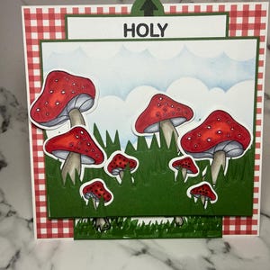 May include: A handmade greeting card with a playful illustration of red mushrooms with white spots set against a green grassy field and a blue sky. The word "HOLY" is printed at the top. The card has a red and white gingham border.