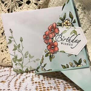 May include: A handmade birthday card with a floral design. The card has a light blue background with a pattern of flowers and leaves. The words "Birthday Wishes" are printed on a white label. An envelope with a floral design is also visible.
