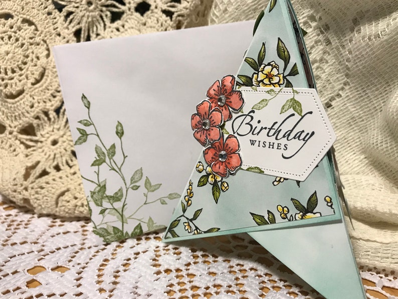 May include: A handmade birthday card with a floral design. The card has a light blue background with a pattern of flowers and leaves. The words "Birthday Wishes" are printed on a white label. An envelope with a floral design is also visible.