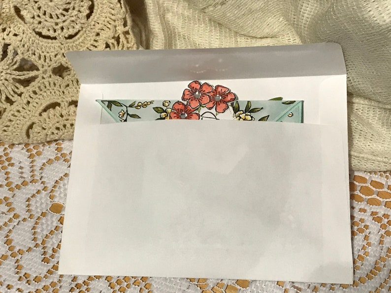 May include: A white envelope containing a floral card. The card has a light blue background with a floral pattern and three coral-coloured flowers. The envelope is partially open, showing the card.