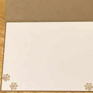 May include: A folded greeting card with a brown exterior and a plain white interior. The bottom left and bottom right corners of the white interior feature small, light brown paw print designs, adding a subtle decorative touch to the card.