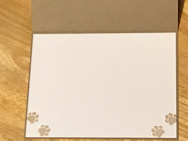 May include: A folded greeting card with a brown exterior and a plain white interior. The bottom left and bottom right corners of the white interior feature small, light brown paw print designs, adding a subtle decorative touch to the card.