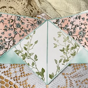 May include: A handmade greeting card with a unique design. The card features a soft pink floral pattern with white flowers and green leaves. The interior has a white background with green leaf accents and a light green border.