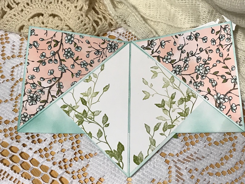 May include: A handmade greeting card with a unique design. The card features a soft pink floral pattern with white flowers and green leaves. The interior has a white background with green leaf accents and a light green border.