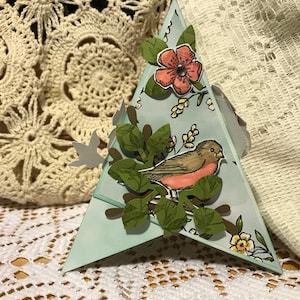 May include: A triangular paper craft with a hand-drawn bird on a branch with green leaves and a pink flower. The craft is light blue with floral accents and a grey ribbon.