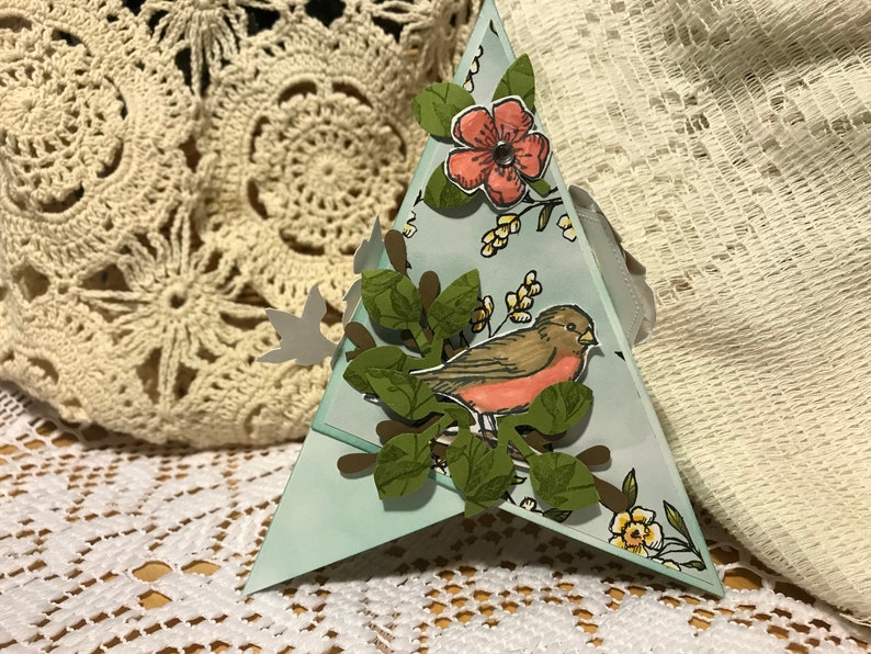 May include: A triangular paper craft with a hand-drawn bird on a branch with green leaves and a pink flower. The craft is light blue with floral accents and a grey ribbon.