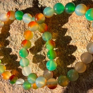 May include: Two circular bracelets made of smooth, polished beads in translucent shades of green, orange, and white. The beads exhibit a natural, marbled appearance with varying colour gradients, strung together to form flexible, continuous loops.