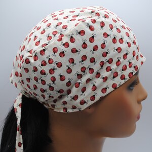 May include: A white fabric head covering with a red ladybug print. The head covering has a long tie that can be tied at the back of the neck.