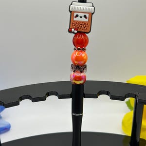 May include: A black pen with a decorative top featuring a cute boba tea design. The pen's shaft is adorned with orange and red beads, and a small bow. The pen is displayed on a black stand.