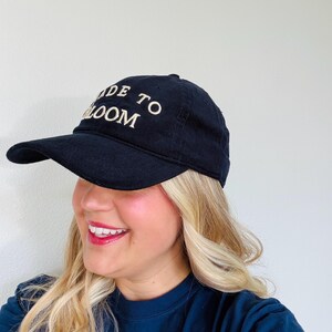 May include: A black corduroy baseball cap featuring the embroidered text MADE TO BLOOM in white capital letters on the front panel. The cap has a curved brim and a structured crown, constructed from a textured fabric with visible vertical ridges.