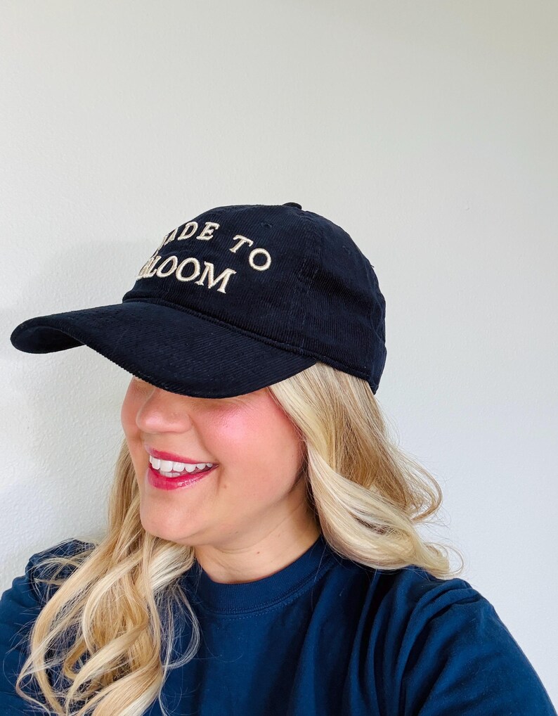 May include: A black corduroy baseball cap featuring the embroidered text MADE TO BLOOM in white capital letters on the front panel. The cap has a curved brim and a structured crown, constructed from a textured fabric with visible vertical ridges.