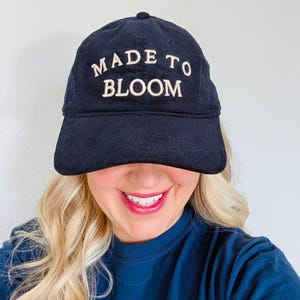 May include: A navy blue corduroy baseball cap featuring the text MADE TO BLOOM embroidered in white thread across the front panel. The cap has a structured crown, a curved brim, and a button at the top center, showcasing a classic six-panel design.