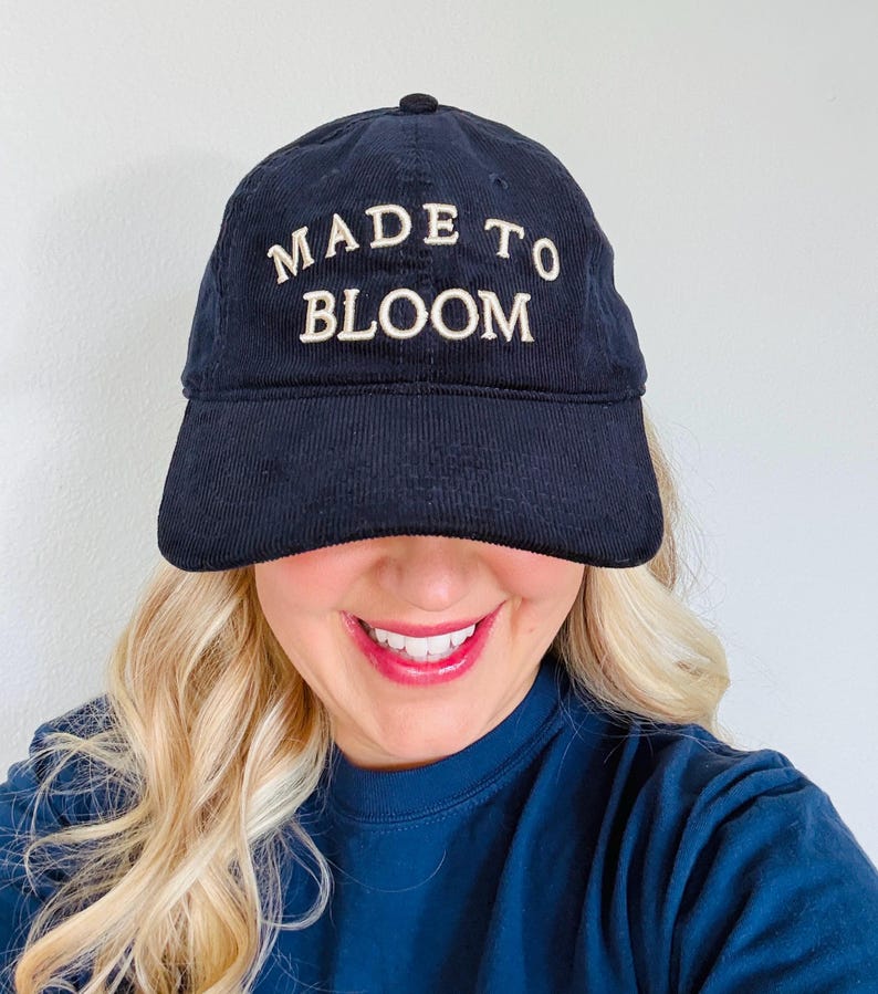 May include: A navy blue corduroy baseball cap featuring the text MADE TO BLOOM embroidered in white thread across the front panel. The cap has a structured crown, a curved brim, and a button at the top center, showcasing a classic six-panel design.
