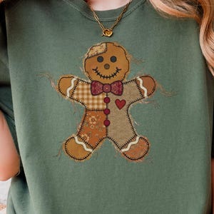 Pode incluir: Uma t-shirt de manga curta verde-azeitona com um gráfico central de um boneco de gengibre. A figura apresenta um design de patchwork em tons de castanho, uma gravata borboleta vermelha, um pequeno coração vermelho e detalhes costurados.