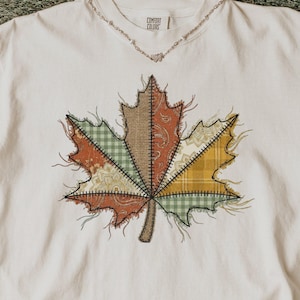 May include: An off-white short-sleeve t-shirt featuring a large, central graphic of a maple leaf composed of various patchwork fabric patterns in shades of brown, green, and yellow, with visible stitched edges and frayed details.