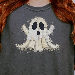 May include: A charcoal gray short-sleeve t-shirt featuring a central graphic of a ghost made from various beige and white fabric patches with frayed edges, black oval eyes, and a black oval mouth, worn over dark green leopard print biker shorts.