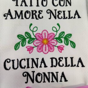 Może przedstawiać: Biały ręcznik kuchenny z haftowanymi r&oacute;żowymi i zielonymi kwiatami i tekstem "Fatto con amore nella cucina della nonna" w kolorze czarnym.