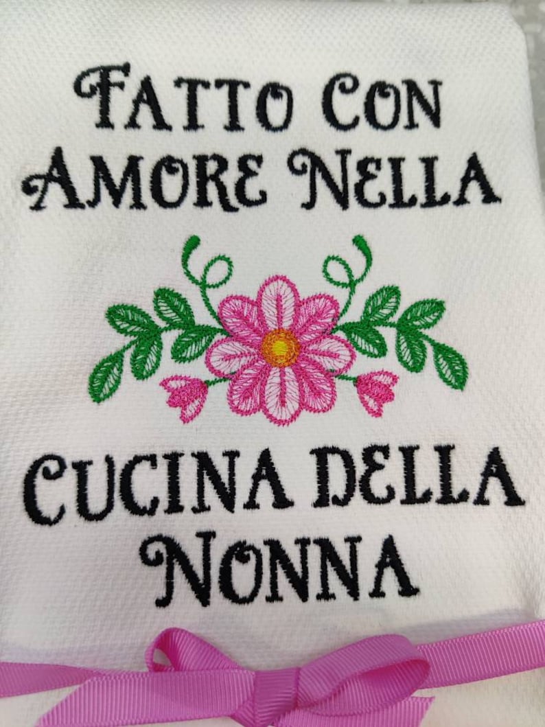 Może przedstawiać: Biały ręcznik kuchenny z haftowanymi r&oacute;żowymi i zielonymi kwiatami i tekstem "Fatto con amore nella cucina della nonna" w kolorze czarnym.