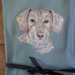 May include: Embroidered gray dog face on a blue kitchen towel with a black ribbon tied around it.