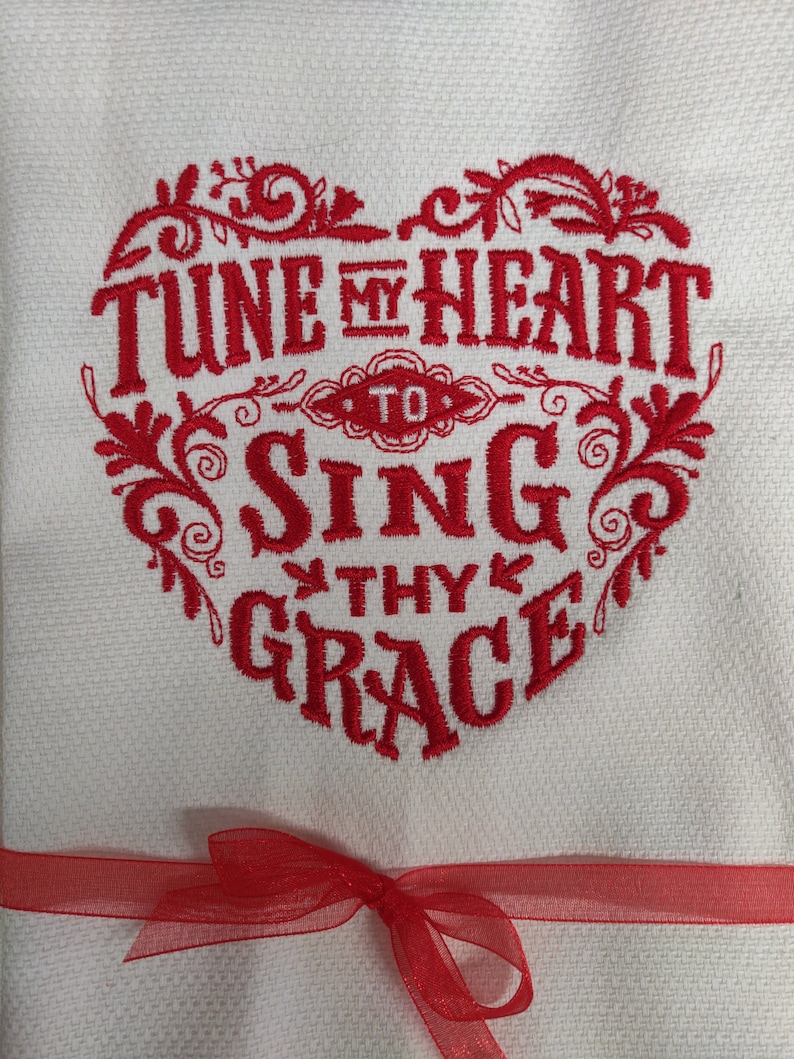 Puede incluir: Toalla de cocina blanca con un coraz&oacute;n bordado en rojo que dice "Tune my heart to sing thy grace". La toalla est&aacute; atada con una cinta roja.