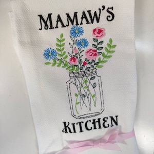 May include: White kitchen towel with black embroidery that reads "Mamaw's Kitchen". The embroidery also includes a mason jar with flowers in pink, blue, and green.