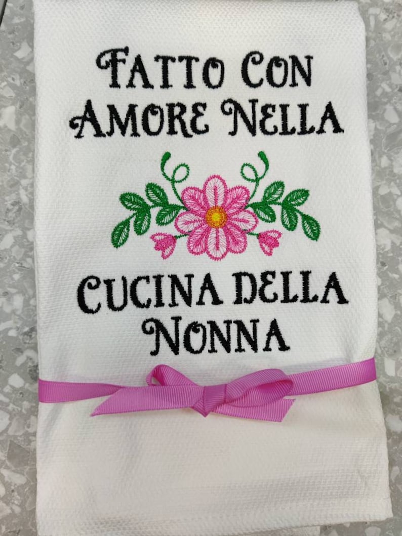 Może przedstawiać: Biały ręcznik kuchenny z r&oacute;żową wstążką zawiązaną wok&oacute;ł. Ręcznik jest haftowany czarnym wzorem z napisem "Fatto con amore nella cucina della nonna".
