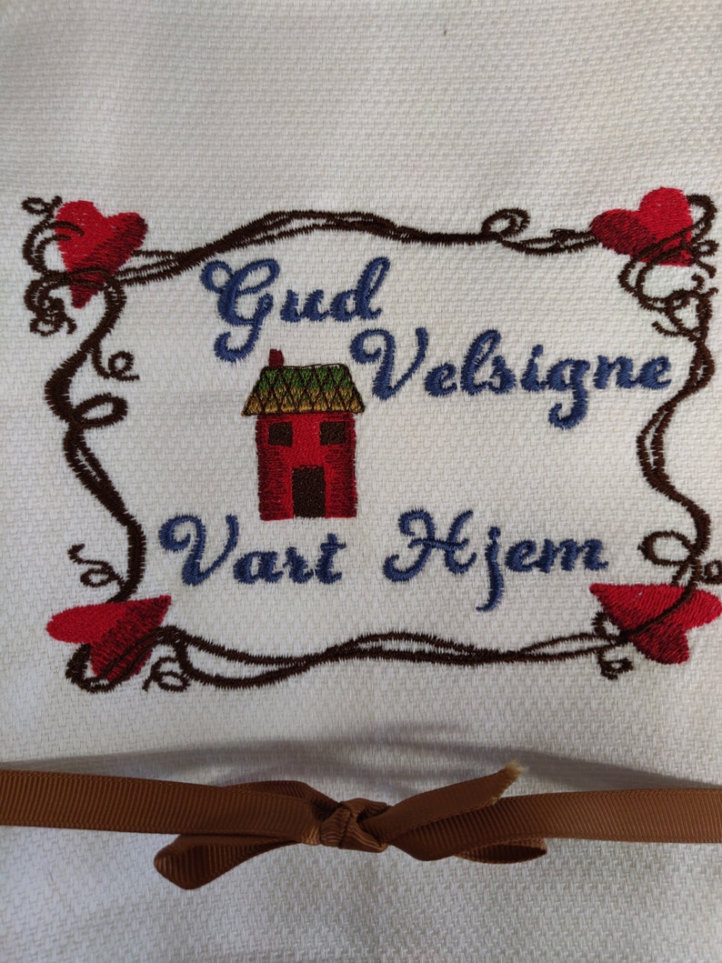 K&ouml;nnte beinhalten: Wei&szlig;es K&uuml;chentuch mit brauner Stickerei eines Hauses, Herzen und dem Text "Gud Velsigne Vart Hjem". Ein braunes Band ist um das Tuch gebunden.