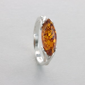 May include: A silver ring featuring a marquise-shaped amber gemstone. The amber is a warm honey colour with inclusions. The ring has a delicate, vine-like design.