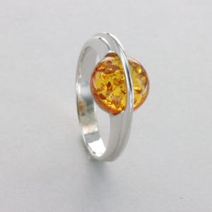 May include: A silver ring with a round amber gemstone set in a bezel setting. The ring has a simple, modern design.