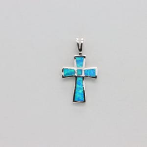 Puede incluir: Colgante de cruz plateada con incrustaciones de ópalo azul-verdoso. La cruz presenta un diseño clásico con una anilla para una cadena. El ópalo tiene una calidad brillante e iridiscente, que añade profundidad e interés visual a la joya.
