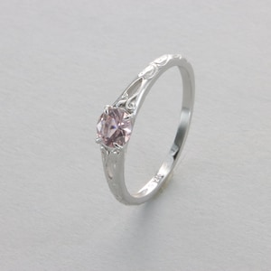 Puede incluir: Un delicado anillo de plata con un diseño floral delicado y una piedra preciosa rosa en el centro.