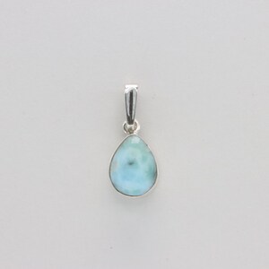 May include: A teardrop-shaped pendant with a light blue gemstone, set in a silver metal frame. The gemstone has a marbled appearance. The pendant has a silver bail for attaching to a chain. Jewelry.