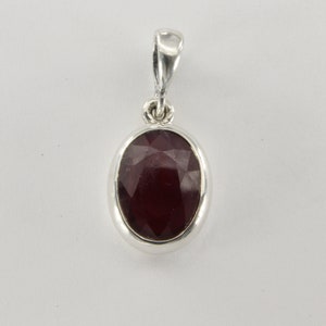 May include: A silver pendant with an oval, faceted, deep red gemstone. The gemstone is set in a polished silver bezel and has a loop for a chain. The pendant is displayed against a white background.