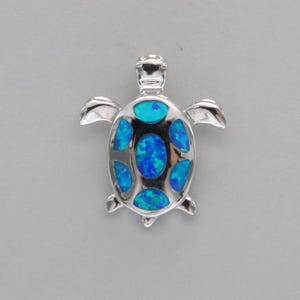 Puede incluir: Un colgante de plata en forma de tortuga con incrustaciones de ópalo azul. La tortuga está mirando hacia adelante con la cabeza levantada y las patas extendidas. La incrustación de ópalo tiene la forma del caparazón de la tortuga y es de un color azul vibrante.