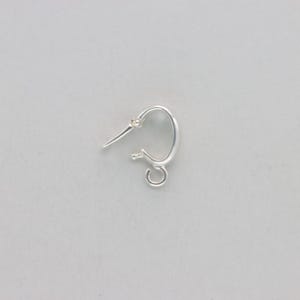 May include: A single, small, silver-colored hoop earring. The earring has a curved design with a post for insertion into the ear and a small loop at the bottom for attaching charms or other decorative elements. The earring is shown against a plain, light background.