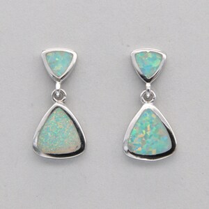 May include: A pair of silver dangle earrings with two teardrop-shaped opals. The top opal is a small triangle and the bottom opal is larger and pear-shaped. The opals are a light green color with white flecks.