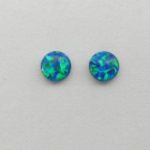 May include: Two round, iridescent opal cabochons. The opals display a mix of blue and green colours, with a speckled pattern. The cabochons are set against a plain, light grey background.