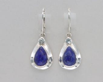 May include: A pair of silver earrings featuring a teardrop-shaped, deep blue lapis lazuli stone. Each earring has a small, round, light blue gemstone above the lapis lazuli, and a silver hook closure.