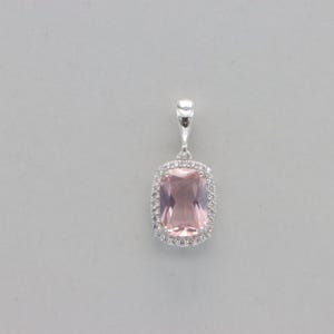 Puede incluir: Un colgante de plata con una gran piedra preciosa rectangular de color rosa claro rodeada por un halo de piedras preciosas más pequeñas y transparentes.