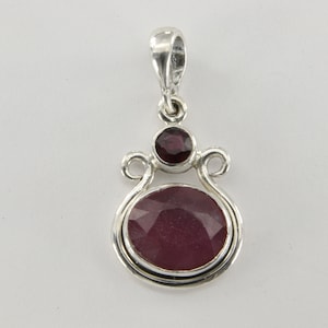 May include: A silver pendant with a large oval-shaped ruby gemstone and a smaller round ruby gemstone. The pendant has a delicate, ornate design.